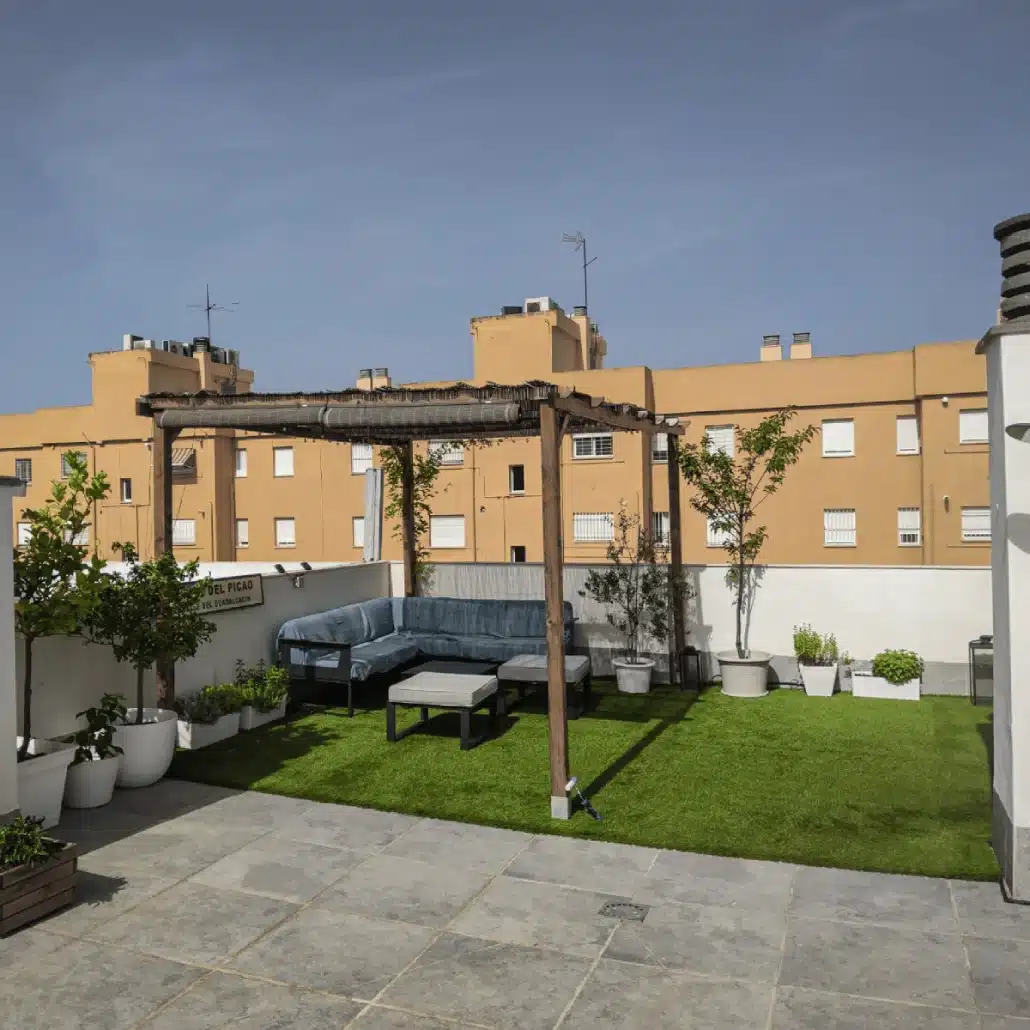 Una azotea con Césped artificial ADRIÁTICO 30mm, plantas en macetas, una pérgola de madera, un sofá seccional azul de exterior y una mesita. Al fondo aparecen edificios de apartamentos amarillos bajo un cielo despejado.